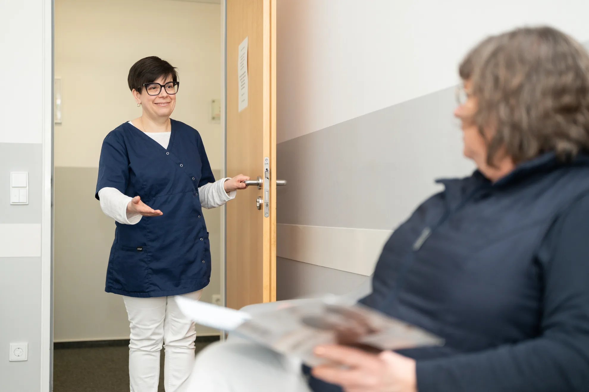 Patientin wird von der Mitarbeiterin lächelnd aus dem Wartezimmer aufgerufen